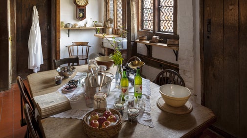 A table in the kitchen at Brockhampton, with bottles of juice, cooking ingredients and equipment, a recipe book, and a basket of apples.